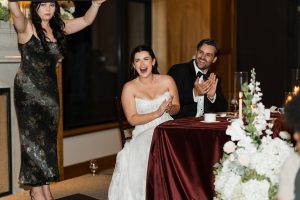 A bride and groom sit at a table, smiling and clapping, as a woman in a black dress raises her arms. Flowers and candles add elegance—perfect inspiration for wedding photographers Toronto recommend.