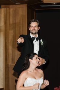 A man in a tuxedo points and smiles behind a laughing woman in a white strapless dress, captured indoors with wooden walls—an authentic moment by Wedding Photographers Toronto.