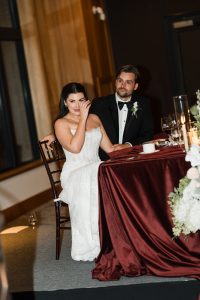 A bride in a white gown wipes a tear while sitting next to the groom at a table with a red tablecloth and floral arrangements, beautifully captured by Wedding Photographers Toronto during the reception.