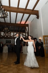 A bride and groom dance together in a formal hall as guests watch. The bride’s white gown and the groom’s black tuxedo are beautifully captured by Wedding Photographers Toronto amid wooden beams, chandeliers, and parquet floors.