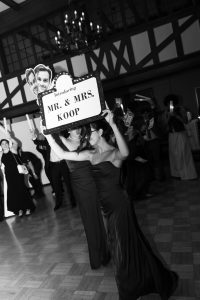 Two women in formal dresses carry a large sign reading Introducing MR. & MRS. KOOP with photos of a couple at an indoor event, captured beautifully in black and white by top Wedding Photographers Toronto.