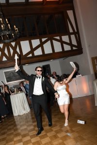 A man in a tuxedo and sunglasses and a woman in a short white dress dance energetically at a formal event as Wedding Photographers Toronto capture guests waving napkins and money scattered on the floor.