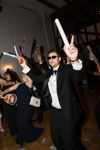 People in formal attire dance at a party, holding glow sticks and cutout face masks. Wedding Photographers Toronto capture a lively scene as dollar bills scatter and a man in sunglasses raises his arms in celebration.