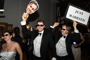 Three people in formal wear and sunglasses celebrate indoors; two men hold a JUST MARRIED sign and a large face cutout, while a woman in a white dress smiles for Wedding Photographers Toronto.