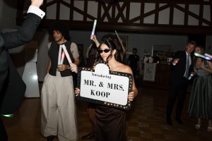 A woman in sunglasses holds a lit sign reading Introducing MR. & MRS. KOOP at a lively indoor party, with people dancing and waving glow sticks—captured perfectly by Wedding Photographers Toronto.