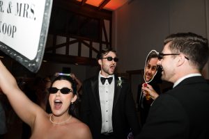 People in formal wear celebrate indoors, wearing sunglasses. One person holds a large face cutout while another raises a “MR. & MRS.” sign—captured brilliantly by Wedding Photographers Toronto.