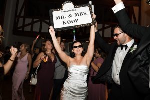 A bride in a white dress and sunglasses holds a sign reading Introducing Mr. & Mrs. Koop, as guests cheer and dance—perfectly captured by top Wedding Photographers Toronto at this lively indoor celebration.