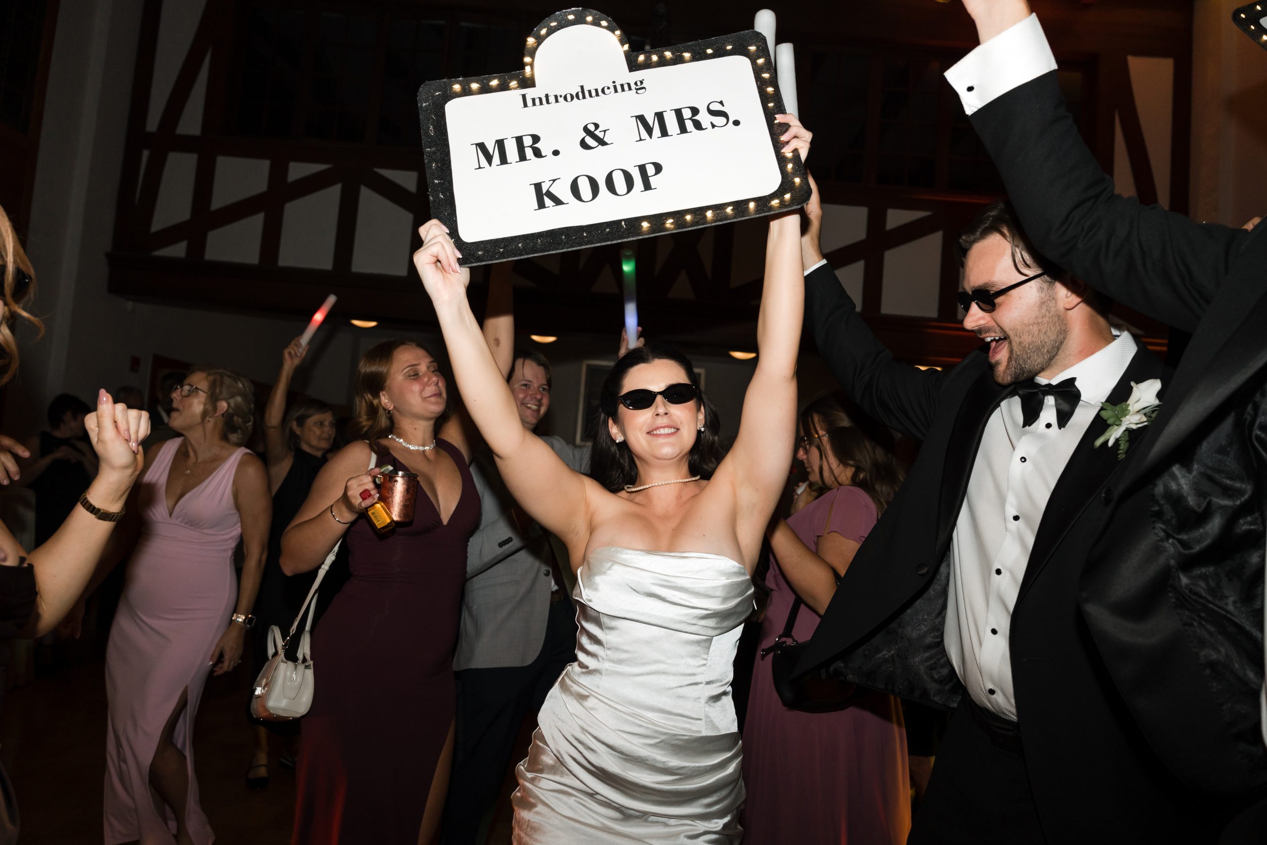 A bride in a white dress and sunglasses holds a sign reading Introducing Mr. & Mrs. Koop, as guests cheer and dance—perfectly captured by top Wedding Photographers Toronto at this lively indoor celebration.