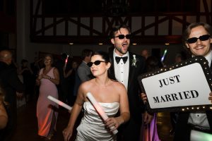 Bride and groom in sunglasses dance at their wedding reception, holding glow sticks. A guest beside them holds a “JUST MARRIED” sign. Wedding Photographers Toronto capture the joyful celebration as guests dance in the background.