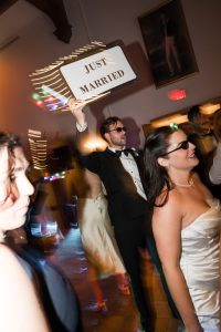 A groom in a tuxedo holds a Just Married sign while walking with the bride and guests at a lively wedding reception, all wearing sunglasses—captured perfectly by top Wedding Photographers Toronto.