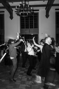 A group of people in formal attire dance energetically under a chandelier, waving glow sticks. Wedding Photographers Toronto capture fake money scattered on the wooden floor and the lively, celebratory atmosphere.
