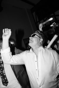 A man in a white shirt and sunglasses dances enthusiastically at a party, his head tilted back and one arm raised—a lively moment often captured by top Wedding Photographers Toronto. The image is in black and white.
