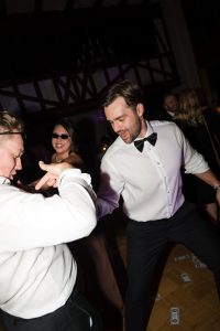 Two men in tuxedos dance energetically at a party, with dollar bills scattered on the floor while a woman in sunglasses smiles in the background—a lively scene captured by Wedding Photographers Toronto.