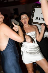 A woman in a white dress dances enthusiastically at a party, holding a lit-up Just Married sign—captured perfectly by Wedding Photographers Toronto as another guest in a dark dress joins the celebration.