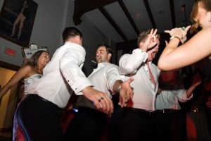 Five people, dressed in formal attire, dance energetically at an indoor party. Captured by expert wedding photographers Toronto, they are smiling and laughing under dim lighting in a lively, joyful atmosphere.
