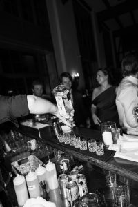 A bartender pours liquor into shot glasses at a busy bar, while people stand and talk in the background. Captured by Wedding Photographers Toronto, the lively scene features drinks and mixers on the counter in black and white.